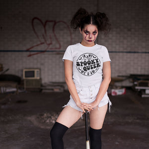 A person with dramatic Halloween makeup, featuring dark eyeshadow and red accents, stands confidently in an abandoned setting, wearing a white t-shirt with the text "In My Spooky Queer Era" printed in a gothic font. The shirt design includes spooky elements like bats and skulls, enhancing the eerie, Halloween theme. The person wears high-waisted, distressed white shorts and black thigh-high socks, holding a baseball bat, adding a rebellious, edgy vibe to the scene. The background features graffiti and old, 