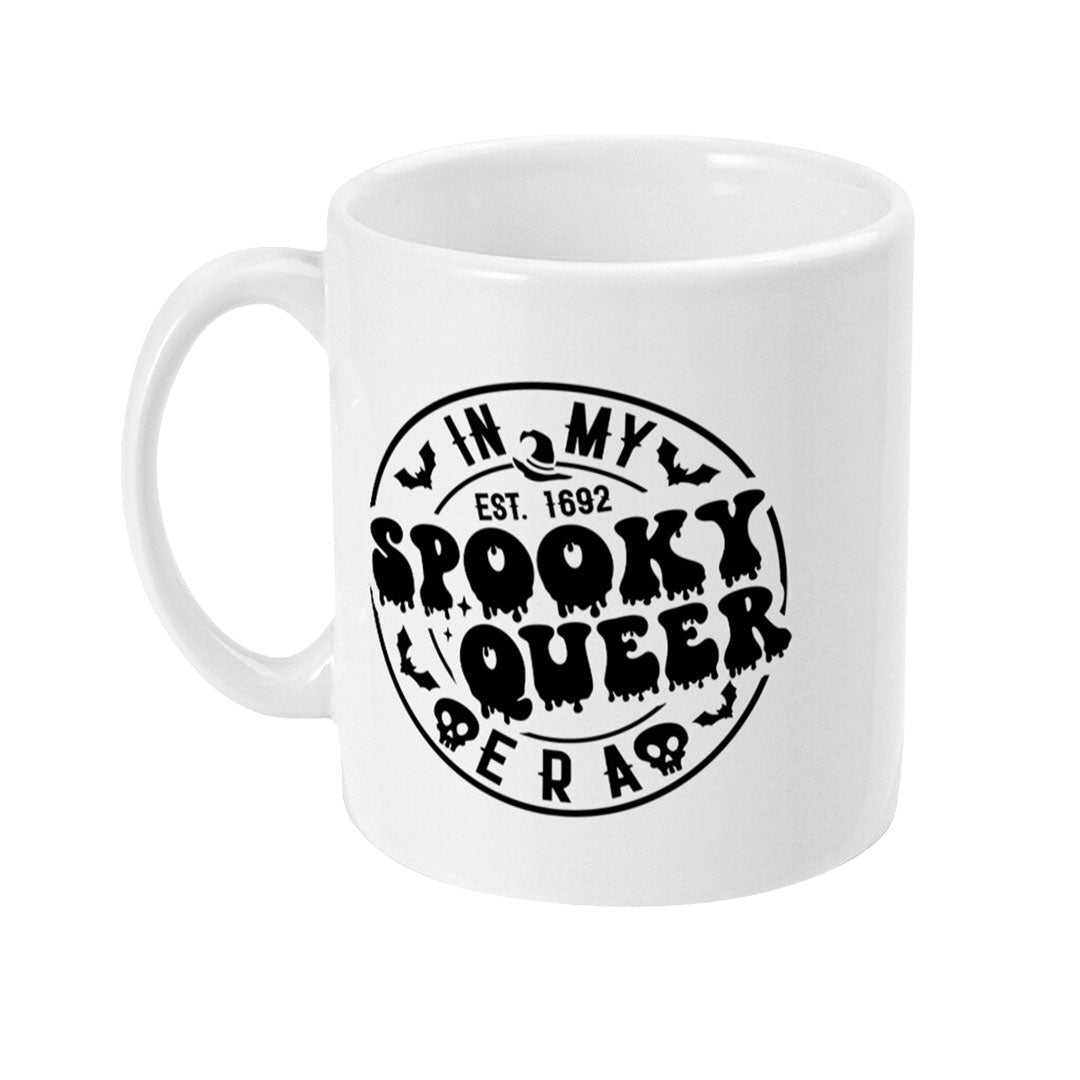 The left side of a white ceramic mug featuring a bold, black design. The design shows a circular emblem with the text "In My Spooky Queer Era Est. 1692" surrounded by bats and skulls, creating a playful yet spooky look.