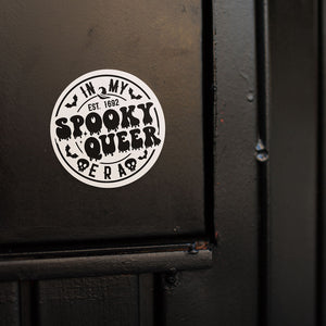 A round white sticker with the text "In My Spooky Queer Era EST. 1692," in black, attached to a black door. The sticker has a dripping letter design with bats and skulls, adding to its eerie and playful Halloween theme.