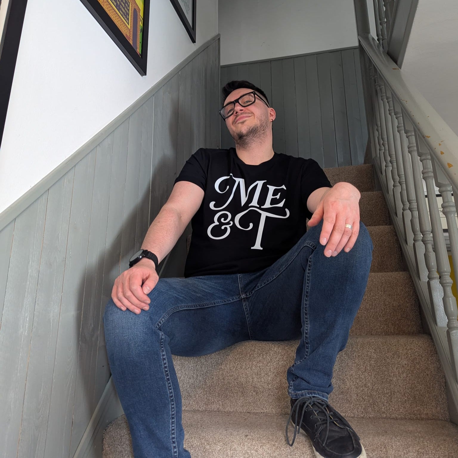 Jamie Lowe sits relaxed on a carpeted staircase with grey panelling, wearing glasses, blue jeans, and a black ‘Me & T’ T-shirt. Jamie wears a size L shirt.