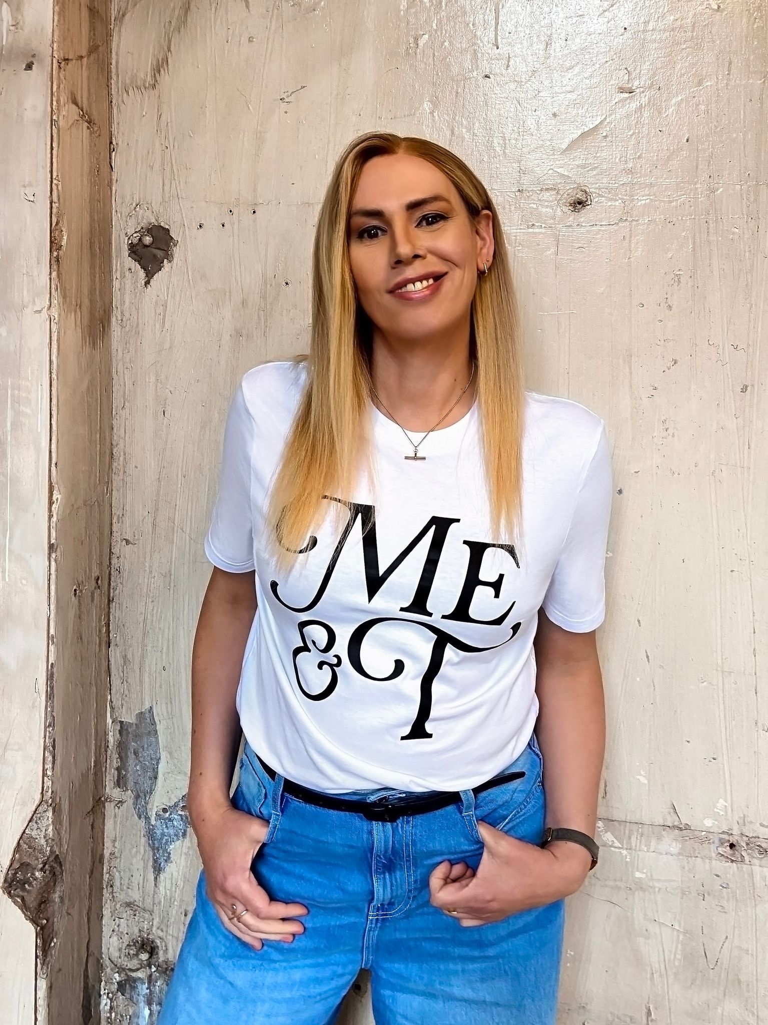 Jodi Fox, a smiling woman with long blonde hair stands against a textured cream wall, wearing a white ‘Me & T’ T-shirt tucked into blue jeans. Jodi wears a size M shirt.