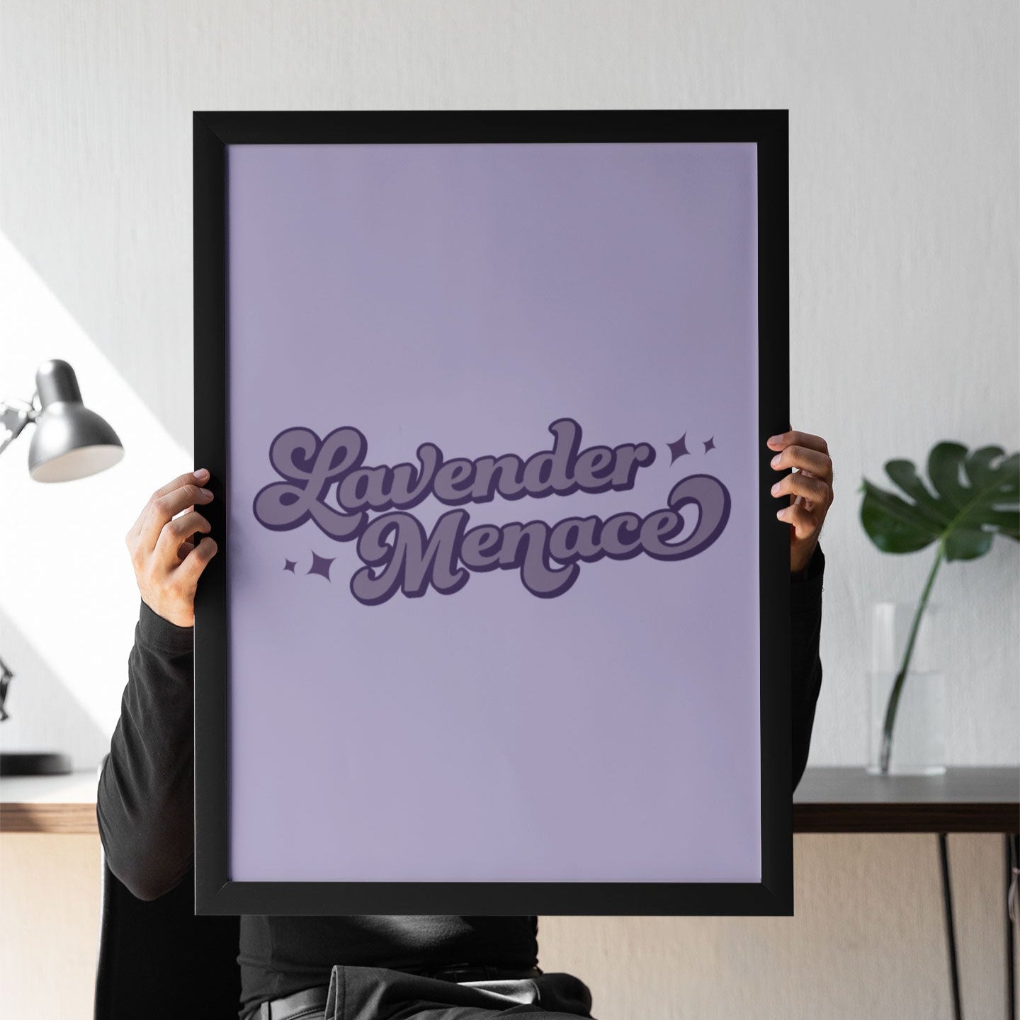 A person sitting at a desk holding up a Lavender Menace print. The stylish purple artwork stands out against a modern, monochrome workspace.