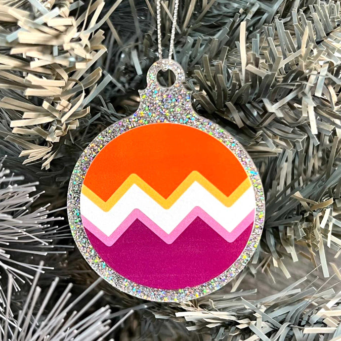 Close up of a white Christmas tree displaying a silver glitter bauble featuring a classic zig zag design in the colours of the lesbian pride flag.