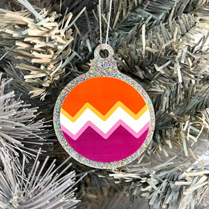 Close up of a white Christmas tree displaying a silver glitter bauble featuring a classic zig zag design in the colours of the lesbian pride flag.