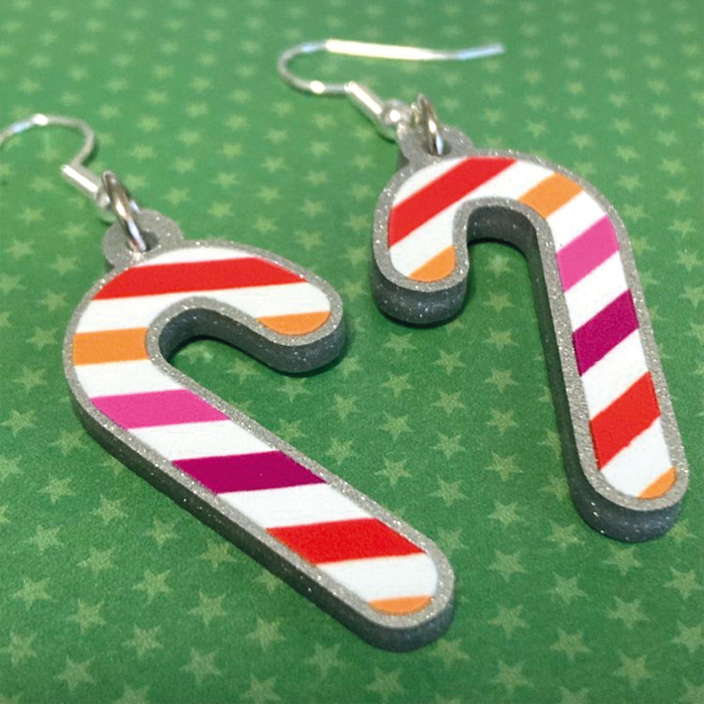 Silver glitter candy cane dangle earrings lay on a green star paper background. The stripes on the candy cane are those of the Lesbian flag; orange, pink, and white. One of the earrings is turned over to show the reverse, which is plain silver glitter.