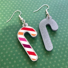 Load image into Gallery viewer, Silver glitter candy cane dangle earrings lay on a green star paper background. The stripes on the candy cane are those of the Lesbian flag; orange, pink, and white. One of the earrings is turned over to show the reverse, which is plain silver glitter.