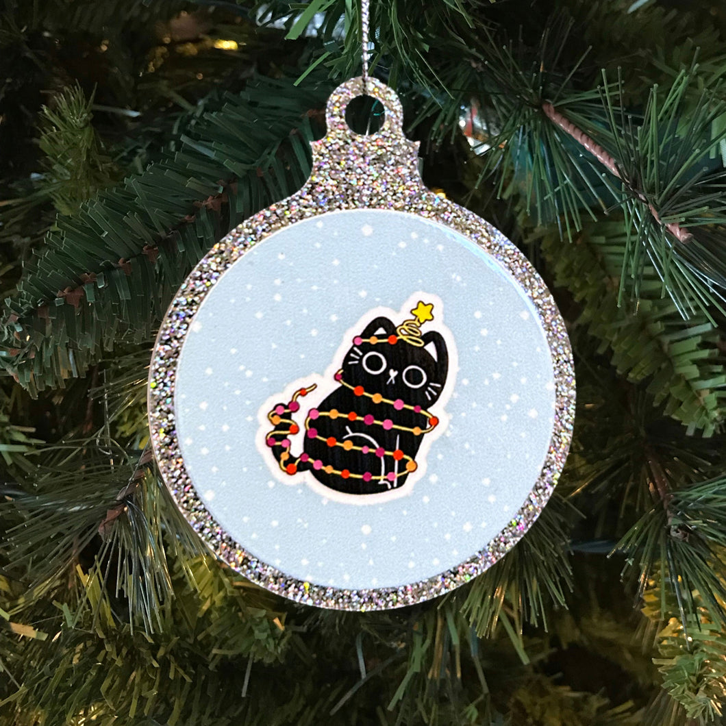 A sparkling Christmas bauble with a black cat tangled in fairy lights in the colours of the lesbian pride flag. The cat sits happily with a star on its head, against a snowy background, hanging on a Christmas tree.