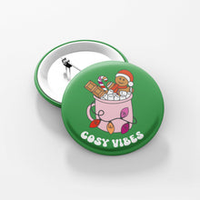 Load image into Gallery viewer, Close up of a green 38mm badge featuring retro text reading &#39;cosy vibes&#39;. The image shows a mug of hot chocolate with marshmallows and a gingerbread man wearing a Santa hat. A candy cane in the mug and lights around the mug are the colours of the lesbian flag.