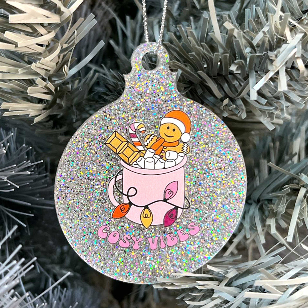 Close up of a white Christmas tree displaying a silver glitter bauble featuring retro text reading 'cosy vibes'. The image shows a mug of hot chocolate with marshmallows and a gingerbread man wearing a Santa hat. A candy cane in the mug and lights around the mug are the colours of the lesbian pride flag.