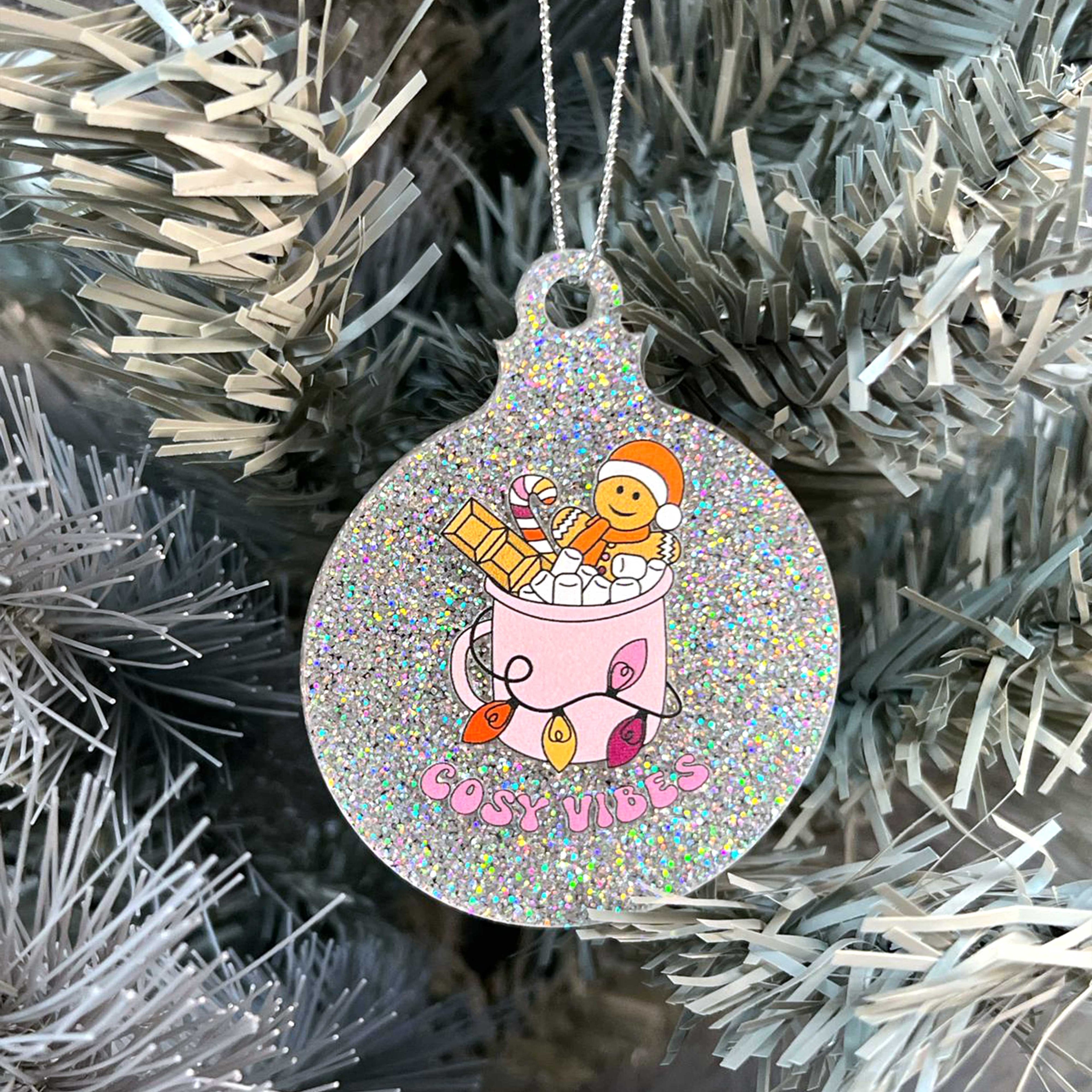 Close up of a white Christmas tree displaying a silver glitter bauble featuring retro text reading 'cosy vibes'. The image shows a mug of hot chocolate with marshmallows and a gingerbread man wearing a Santa hat. A candy cane in the mug and lights around the mug are the colours of the lesbian pride flag.