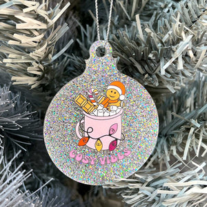 Close up of a white Christmas tree displaying a silver glitter bauble featuring retro text reading 'cosy vibes'. The image shows a mug of hot chocolate with marshmallows and a gingerbread man wearing a Santa hat. A candy cane in the mug and lights around the mug are the colours of the lesbian pride flag.