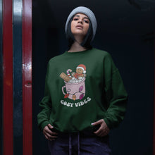 Load image into Gallery viewer, Young woman in a beanie hat wearing a Bottle Green sweatshirt featuring retro text reading &#39;cosy vibes&#39;. The image shows a mug of hot chocolate with marshmallows and a gingerbread man wearing a Santa hat. A candy cane in the mug and lights around the mug are the colours of the lesbian pride flag.