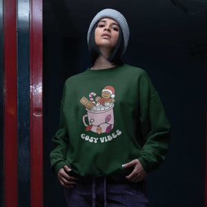 Young woman in a beanie hat wearing a Bottle Green sweatshirt featuring retro text reading 'cosy vibes'. The image shows a mug of hot chocolate with marshmallows and a gingerbread man wearing a Santa hat. A candy cane in the mug and lights around the mug are the colours of the lesbian pride flag.