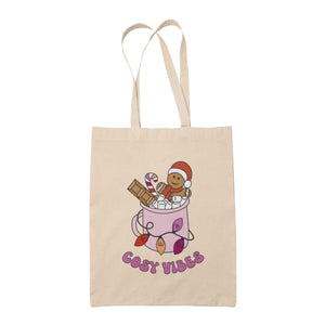 Natural tote bag featuring retro text reading 'cosy vibes'. The image shows a mug of hot chocolate with marshmallows and a gingerbread man wearing a Santa hat. A candy cane in the mug and lights around the mug are the colours of the lesbian pride flag.