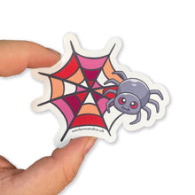 Load image into Gallery viewer, A hand holding a cute spider sticker with a lesbian pride web design. The web is coloured with shades of red, orange, and pink, symbolising the lesbian flag.