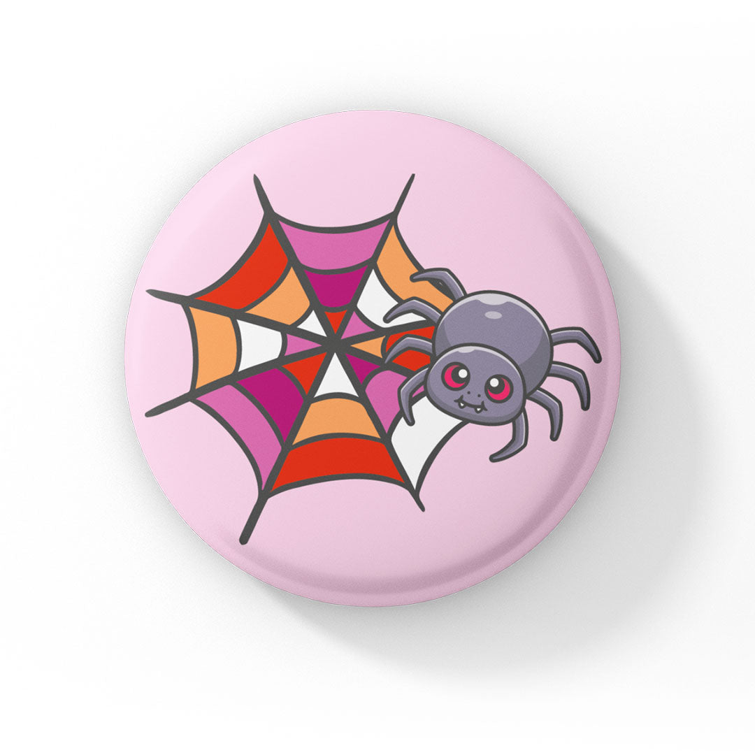 A circular badge featuring a grey spider with pink eyes on a web coloured in red, orange, pink, and white, representing the lesbian pride flag. The badge has a light pink background.