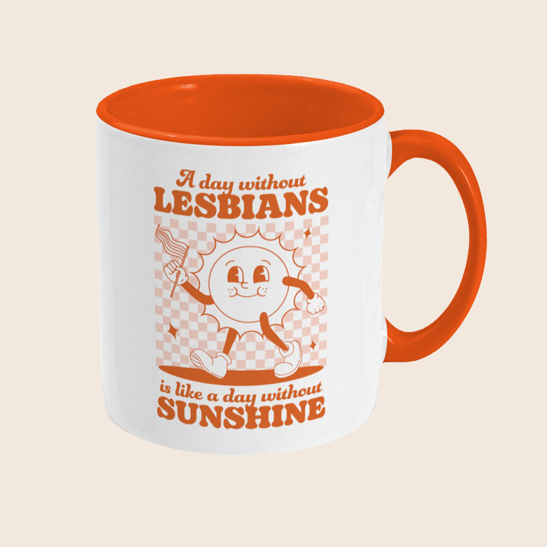 White ceramic mug with an orange interior and handle. The sun character is centered, and the full text is visible: “A day without LESBIANS is like a day without SUNSHINE,” in retro orange lettering with decorative stars and a checked background.