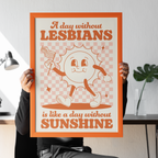 Seated person holding an orange-framed art print in front of their face. The design features a smiling sun holding a flag on a pink chequerboard background, with text reading “A day without lesbians is like a day without sunshine”.