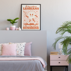 Black-framed “A day without lesbians is like a day without sunshine” art print featuring a smiling sun and pink chequerboard pattern, displayed above a bed with grey bedding and pink cushions, beside plants and ornaments.