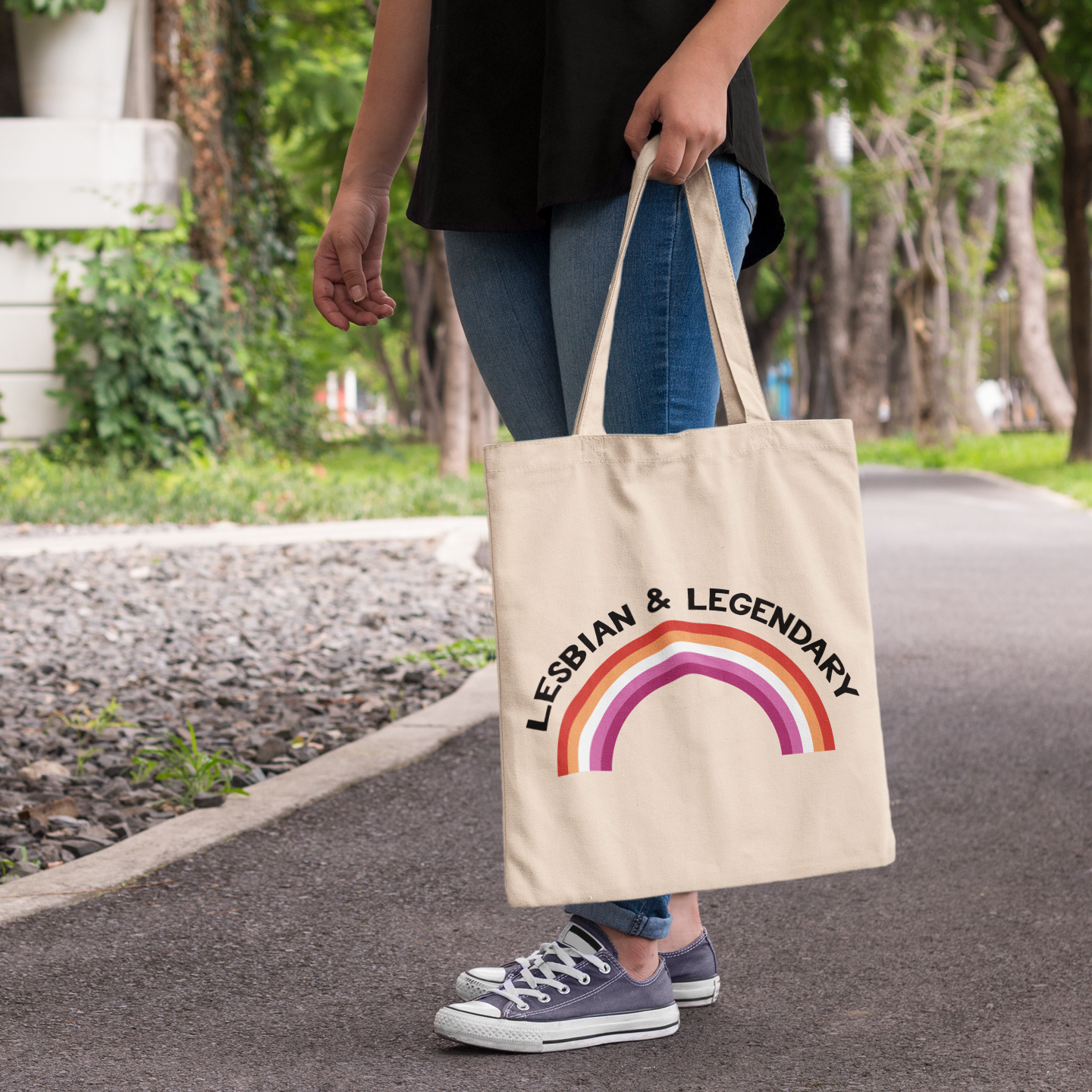 Natural cotton tote bag with “Lesbian & Legendary” text and a lesbian pride rainbow, worn over the shoulder in an outdoor setting.