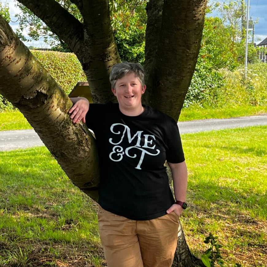 A person stands smiling under a tree in the countryside, wearing a black ‘Me & T’ T-shirt and brown trousers, with one arm resting on a branch.