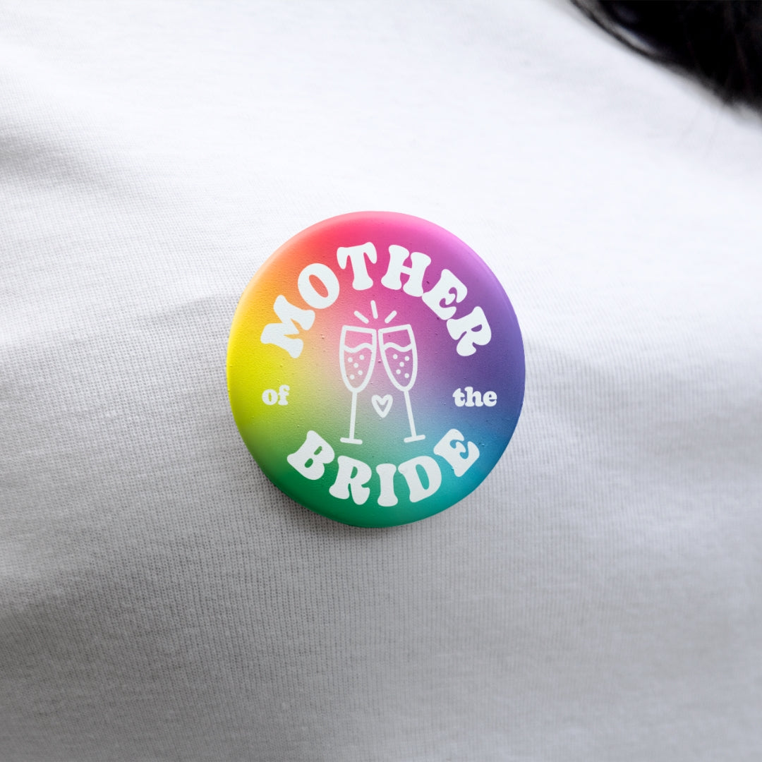 Mother of the Bride 38mm rainbow badge pinned to white fabric, showing champagne glasses and bold white lettering.