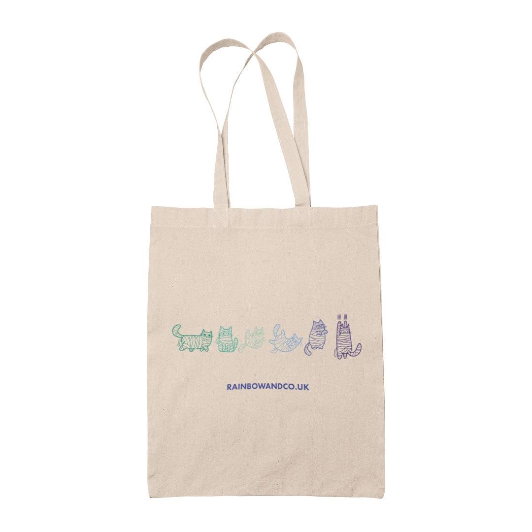 A natural canvas tote bag displaying a line drawing of six mummy cats in shades of green, teal, and blue, representing the colours of the Gay pride flag. Each cat is wrapped like a mummy, with distinctive poses and expressions.