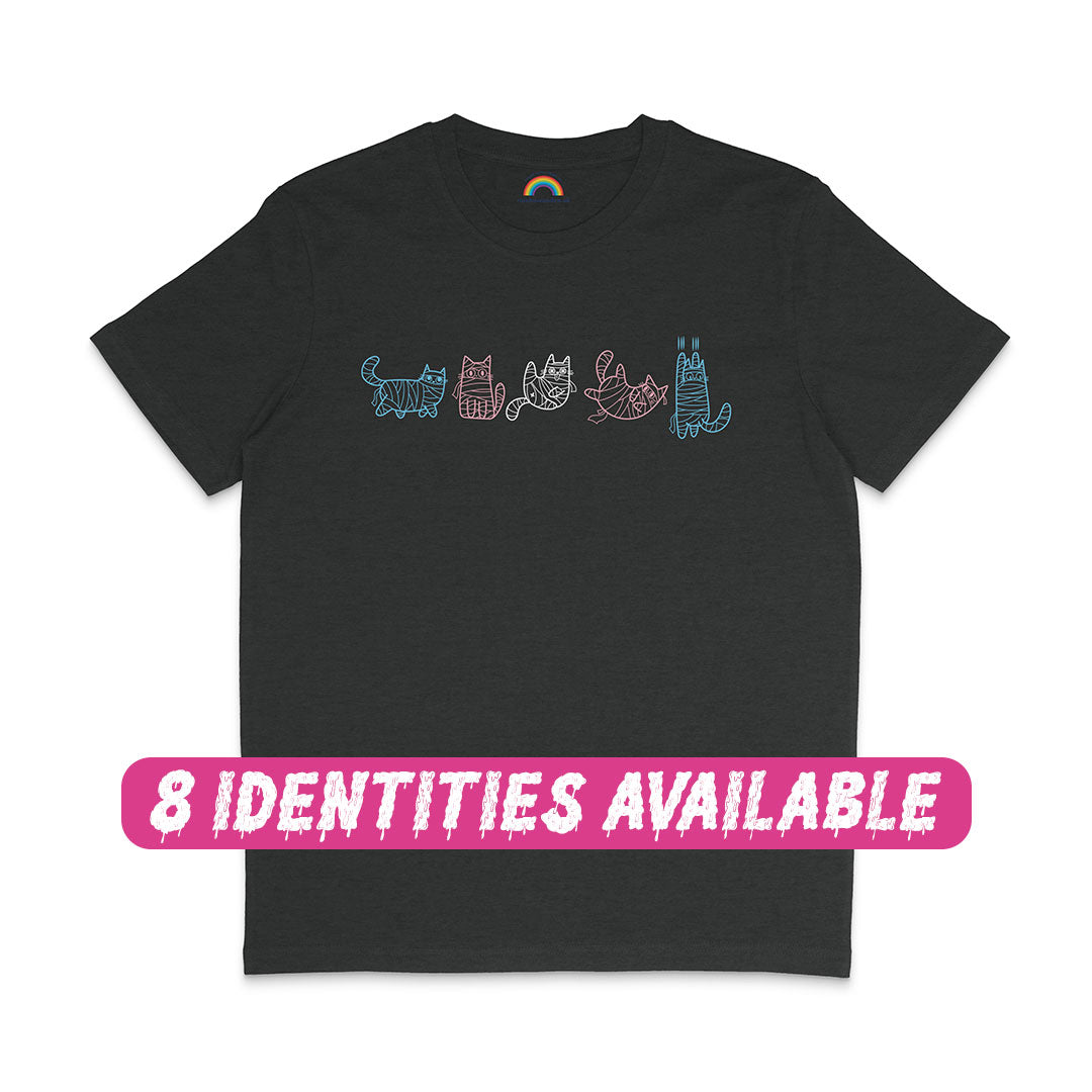 A black T-shirt featuring five mummy cat illustrations in the colours of the transgender flag. The cats are drawn in light blue, pink, and white, each wrapped in bandages and posed uniquely across the chest of the shirt. A pink text overlay at the bottom reads "8 IDENTITIES AVAILABLE," highlighting the diverse options for this design.