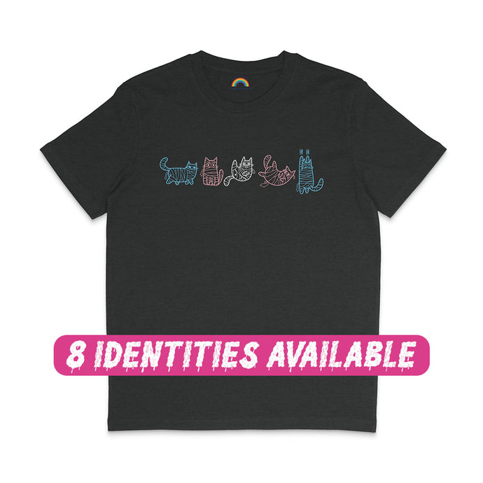 A black T-shirt featuring five mummy cat illustrations in the colours of the transgender flag. The cats are drawn in light blue, pink, and white, each wrapped in bandages and posed uniquely across the chest of the shirt. A pink text overlay at the bottom reads 