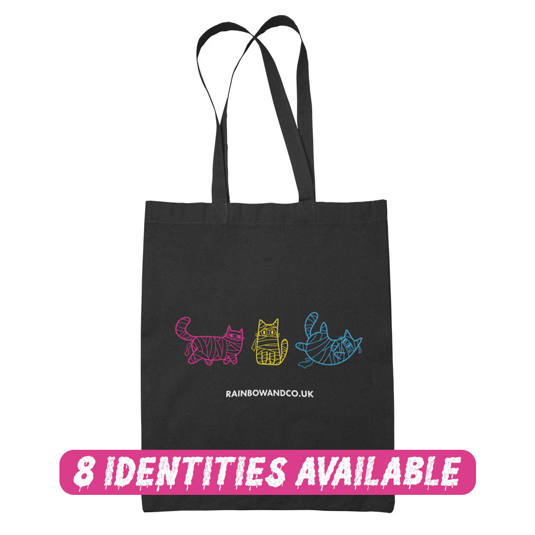 A black tote bag with three mummy cats in different pride colours: pink, yellow, and blue. The bottom of the bag reads "rainbowandco.uk," and a banner across the bottom says "8 Identities Available" in pink lettering.