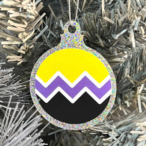 Close up of a white Christmas tree displaying a silver glitter bauble featuring a classic zig zag design in the colours of the non binary pride flag.
