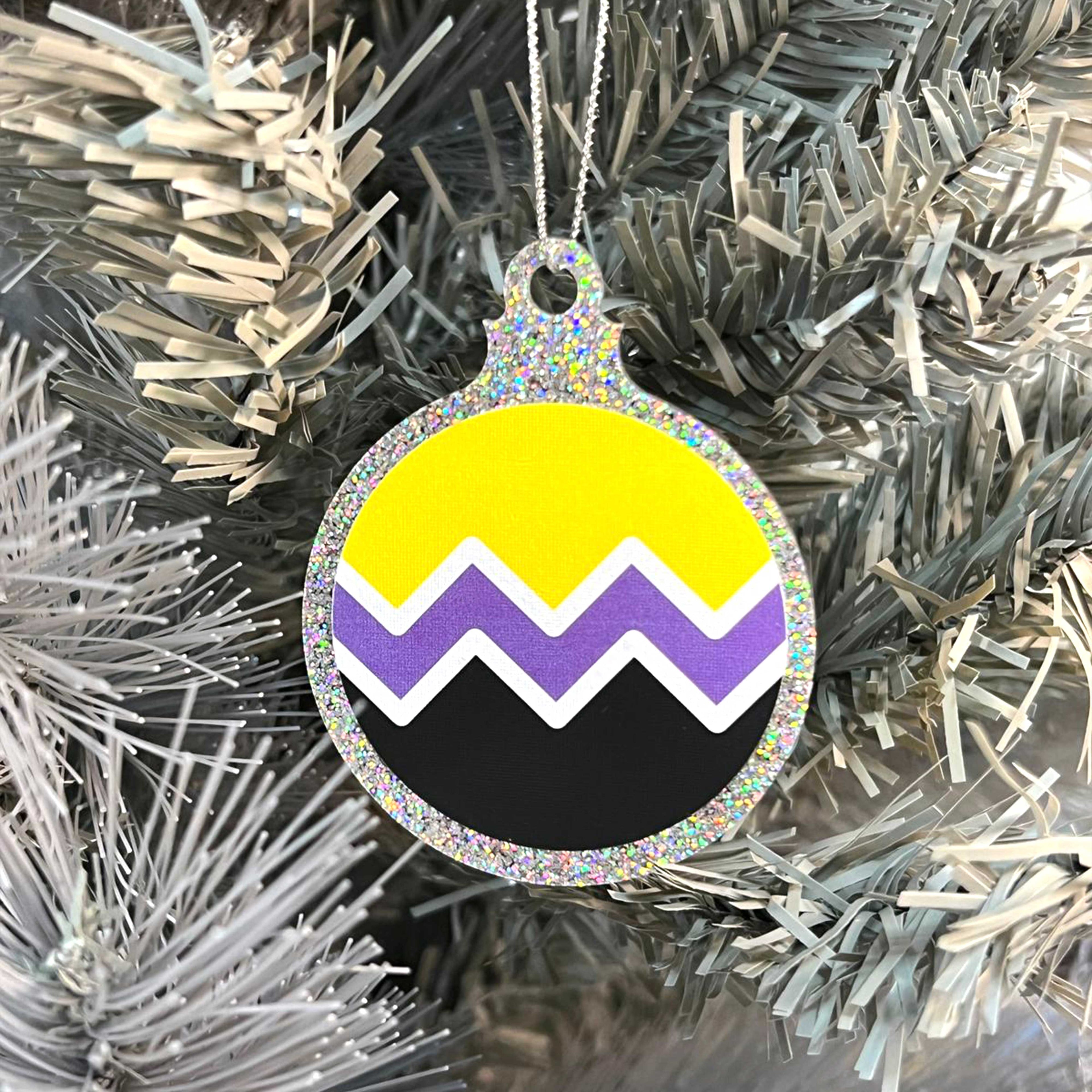 Close up of a white Christmas tree displaying a silver glitter bauble featuring a classic zig zag design in the colours of the non binary pride flag.