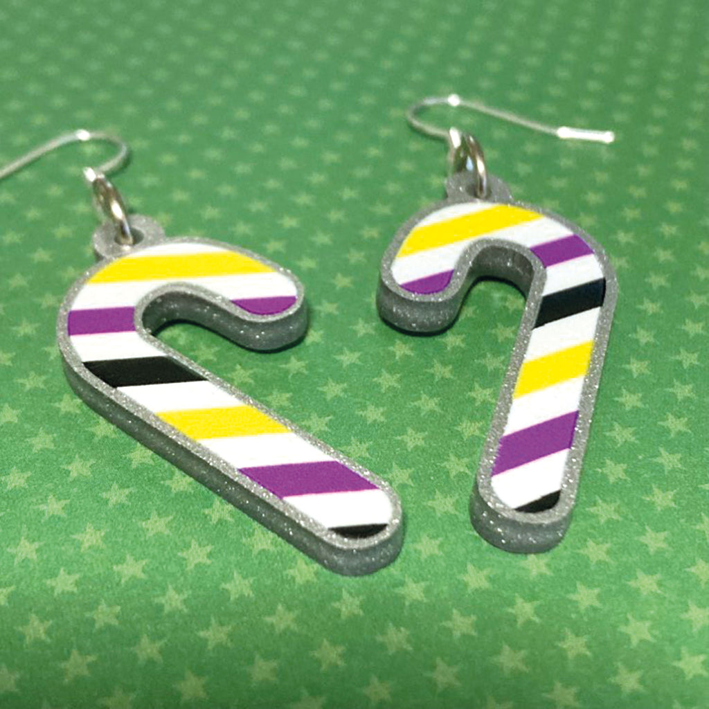 Silver glitter candy cane dangle earrings lay on a green star paper background. The stripes on the candy cane are those of the Non Binary flag; yellow, purple, black, and white.
