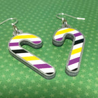 Silver glitter candy cane dangle earrings lay on a green star paper background. The stripes on the candy cane are those of the Non Binary flag; yellow, purple, black, and white.