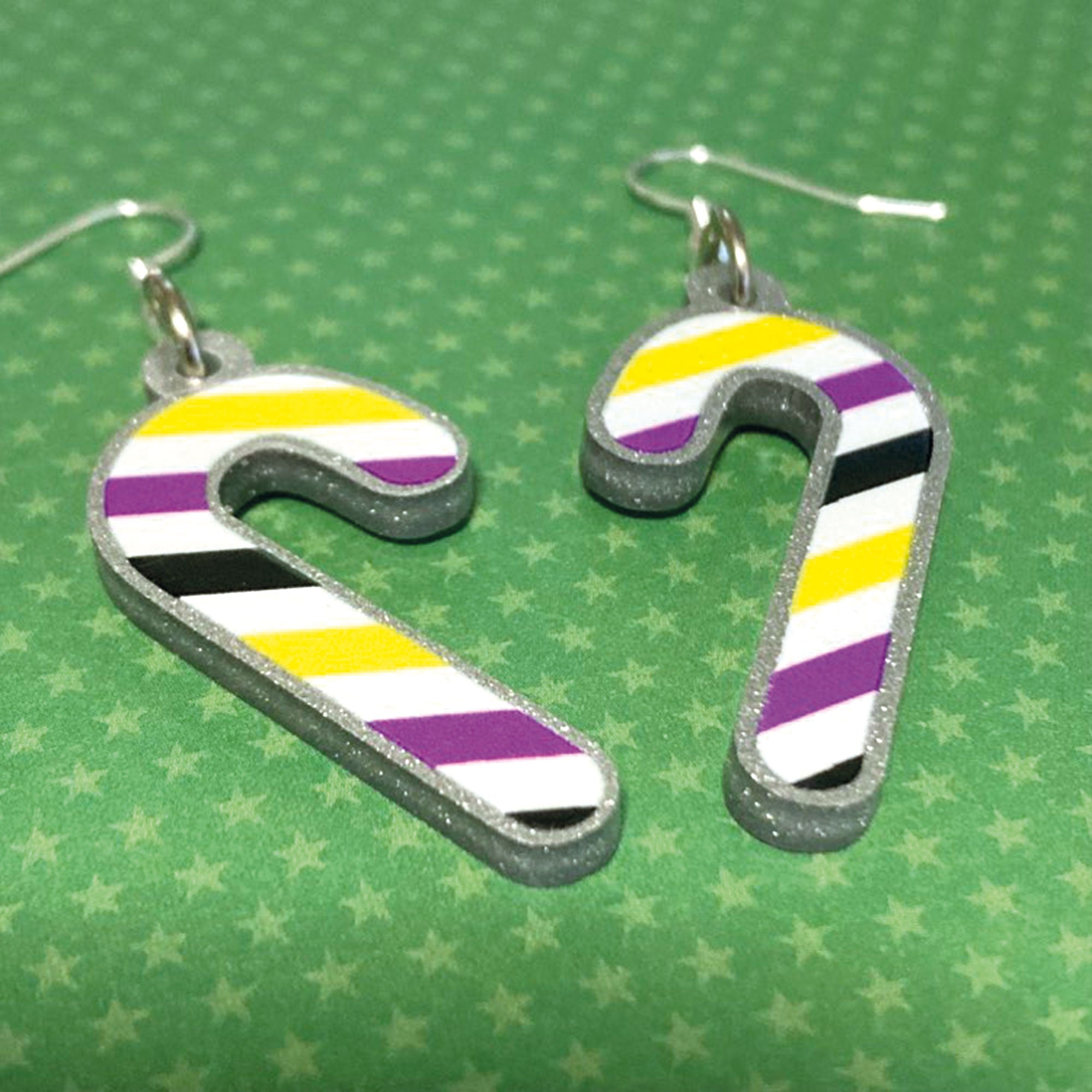 Silver glitter candy cane dangle earrings lay on a green star paper background. The stripes on the candy cane are those of the Non Binary flag; yellow, purple, black, and white.