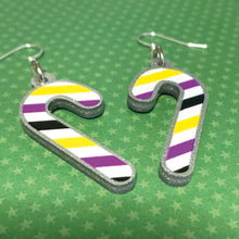 Load image into Gallery viewer, Silver glitter candy cane dangle earrings lay on a green star paper background. The stripes on the candy cane are those of the Non Binary flag; yellow, purple, black, and white.