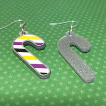 Load image into Gallery viewer, Silver glitter candy cane dangle earrings lay on a green star paper background. The stripes on the candy cane are those of the Non Binary flag; yellow, purple, black, white. One of the earrings is turned over to show the reverse, which is plain silver glitter.