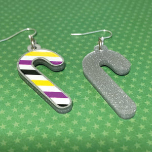 Silver glitter candy cane dangle earrings lay on a green star paper background. The stripes on the candy cane are those of the Non Binary flag; yellow, purple, black, white. One of the earrings is turned over to show the reverse, which is plain silver glitter.