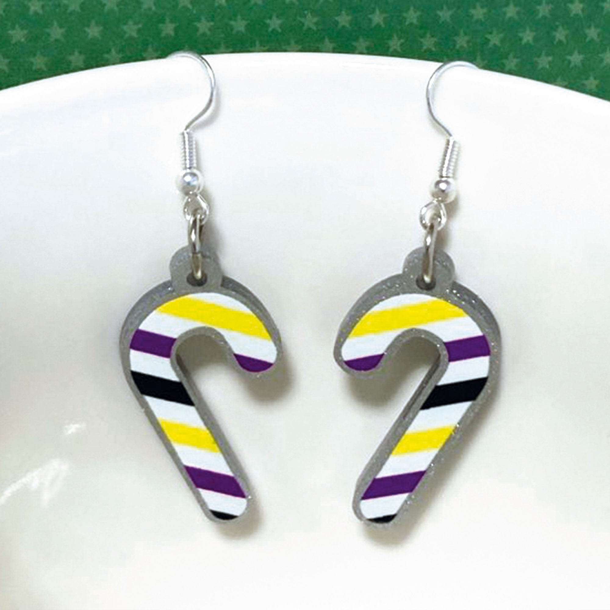 Silver glitter candy cane dangle earrings hung over the edge of a mug. The stripes on the candy cane are those of the Non Binary flag; yellow, purple, black, and white.