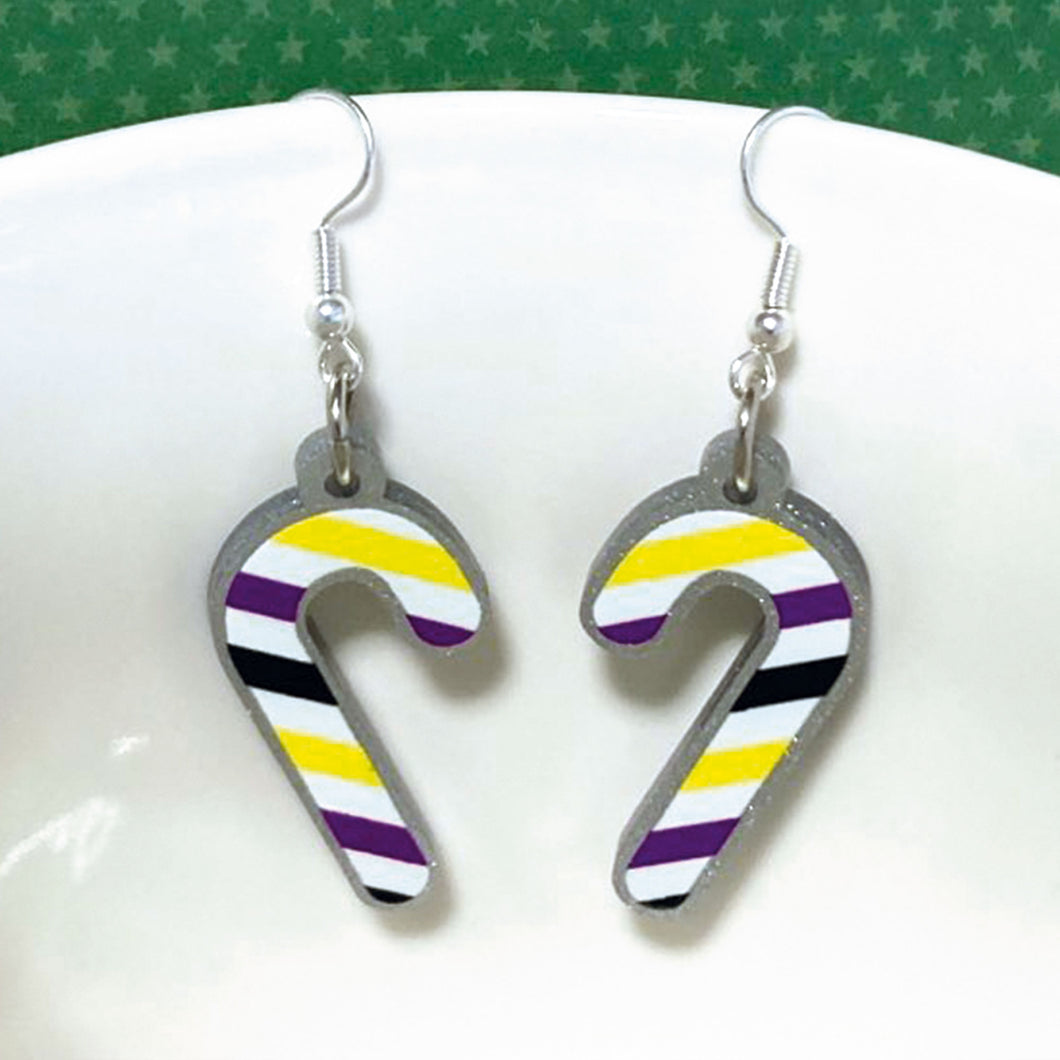Silver glitter candy cane dangle earrings hung over the edge of a mug. The stripes on the candy cane are those of the Non Binary flag; yellow, purple, black, and white.
