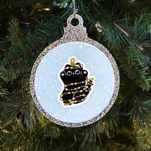 Load image into Gallery viewer,  A glittery Christmas bauble showcasing a black cat wrapped in fairy lights coloured in the non-binary pride flag. The cat has a cute expression and a star atop its head, set against a snowy backdrop on a festive tree.