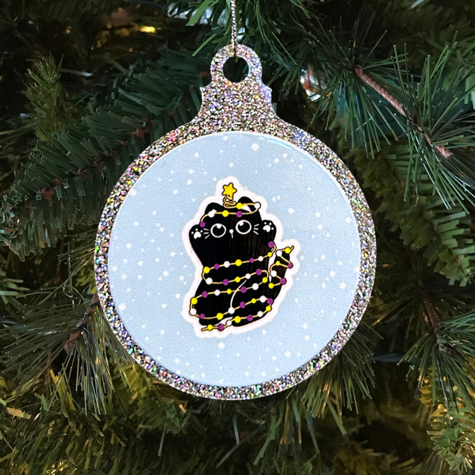  A glittery Christmas bauble showcasing a black cat wrapped in fairy lights coloured in the non-binary pride flag. The cat has a cute expression and a star atop its head, set against a snowy backdrop on a festive tree.