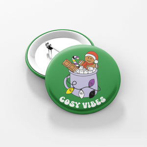Close up of a green 38mm badge featuring retro text reading 'cosy vibes'. The image shows a mug of hot chocolate with marshmallows and a gingerbread man wearing a Santa hat. A candy cane in the mug and lights around the mug are the colours of the non binary flag.