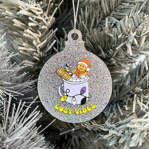 Close up of a white Christmas tree displaying a silver glitter bauble featuring retro text reading 'cosy vibes'. The image shows a mug of hot chocolate with marshmallows and a gingerbread man wearing a Santa hat. A candy cane in the mug and lights around the mug are the colours of the non binary pride flag.