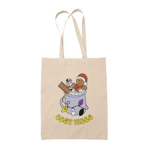 Natural tote bag featuring retro text reading 'cosy vibes'. The image shows a mug of hot chocolate with marshmallows and a gingerbread man wearing a Santa hat. A candy cane in the mug and lights around the mug are the colours of the non binary pride flag.