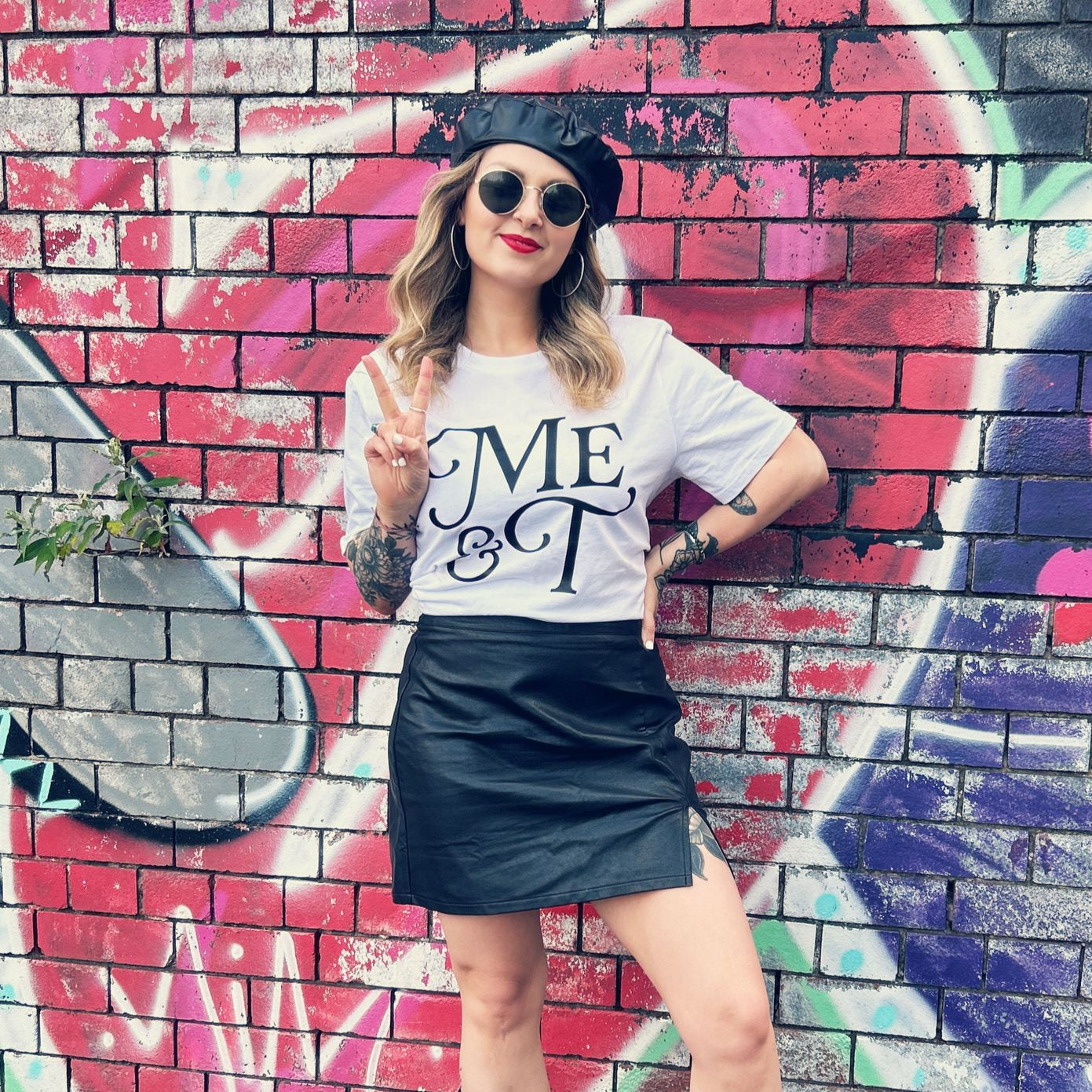 Paige Hughes with wavy blonde hair, wearing a black beret, sunglasses, and a white ‘Me & T’ T-shirt poses with a peace sign in front of a colourful graffiti wall. Paige wears a size L shirt.
