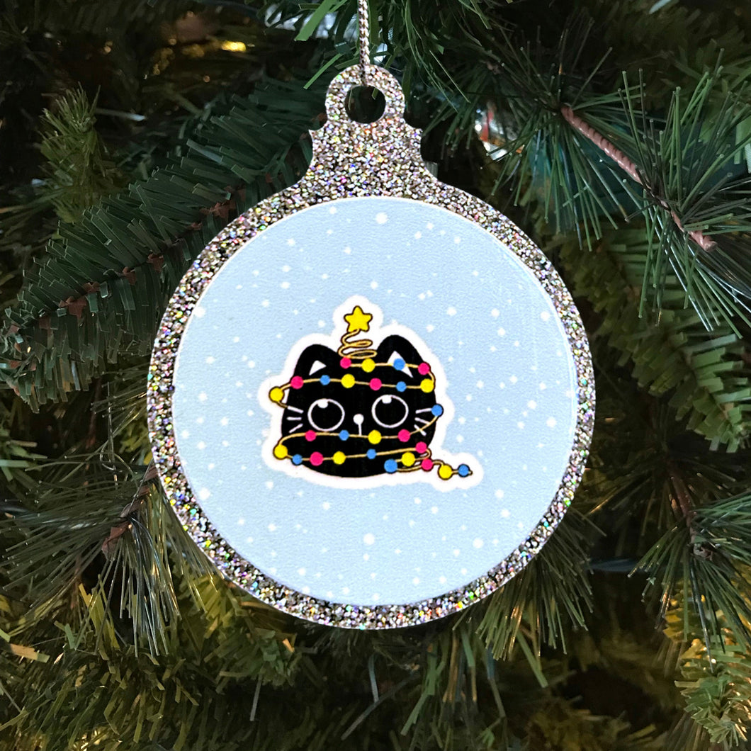 A sparkling Christmas bauble featuring a black cat wrapped in fairy lights in the colours of the pansexual pride flag. The cat sits contentedly with a star on its head against a snowy background, hanging on a Christmas tree.