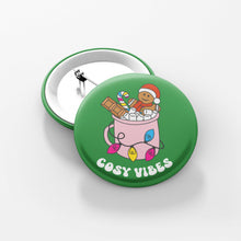Load image into Gallery viewer, Close up of a green 38mm badge featuring retro text reading &#39;cosy vibes&#39;. The image shows a mug of hot chocolate with marshmallows and a gingerbread man wearing a Santa hat. A candy cane in the mug and lights around the mug are the colours of the pansexual flag.