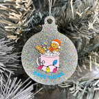 Close up of a white Christmas tree displaying a silver glitter bauble featuring retro text reading 'cosy vibes'. The image shows a mug of hot chocolate with marshmallows and a gingerbread man wearing a Santa hat. A candy cane in the mug and lights around the mug are the colours of the pansexual pride flag.
