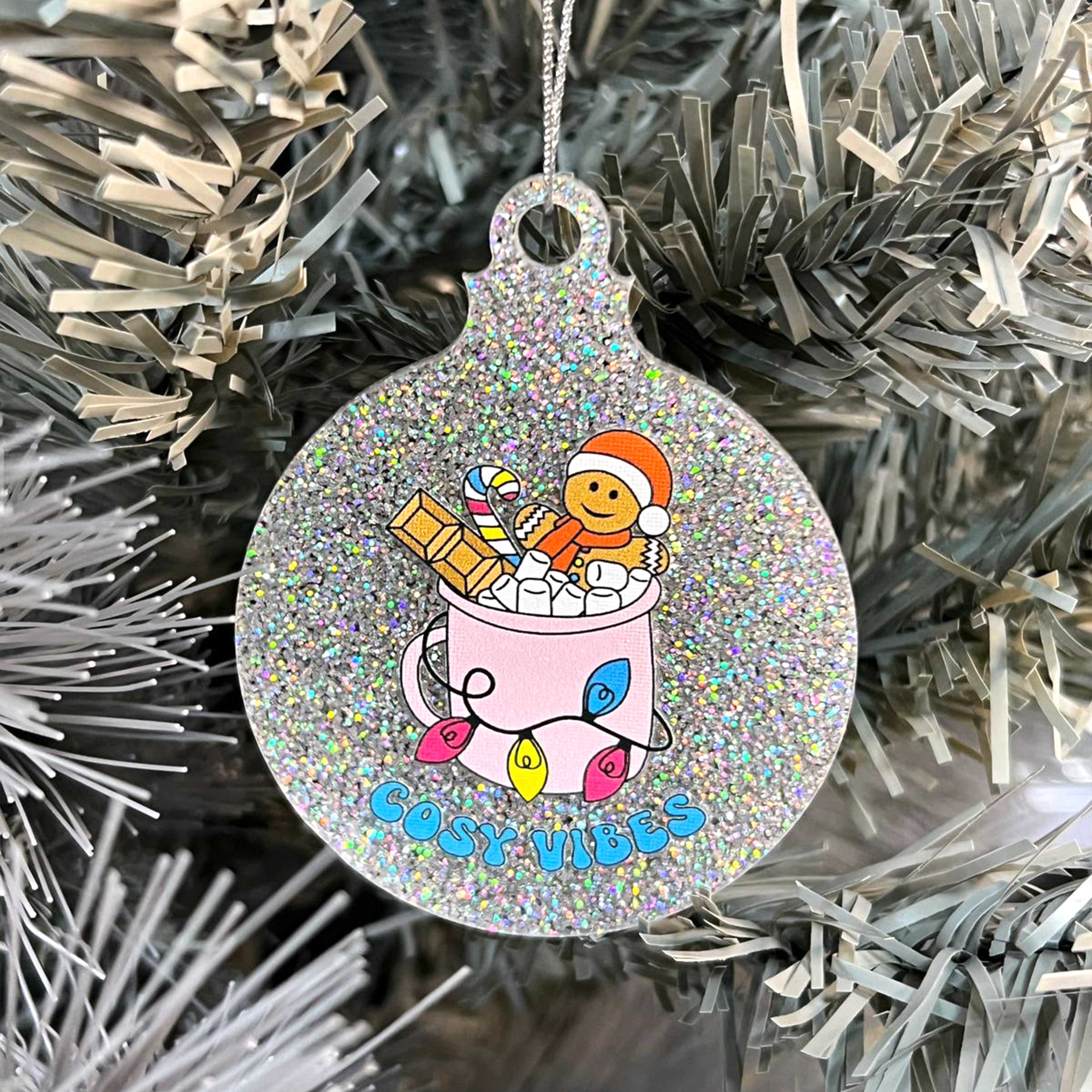 Close up of a white Christmas tree displaying a silver glitter bauble featuring retro text reading 'cosy vibes'. The image shows a mug of hot chocolate with marshmallows and a gingerbread man wearing a Santa hat. A candy cane in the mug and lights around the mug are the colours of the pansexual pride flag.
